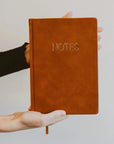 hands holding brown leather journal that says 'NOTES' on it in gold