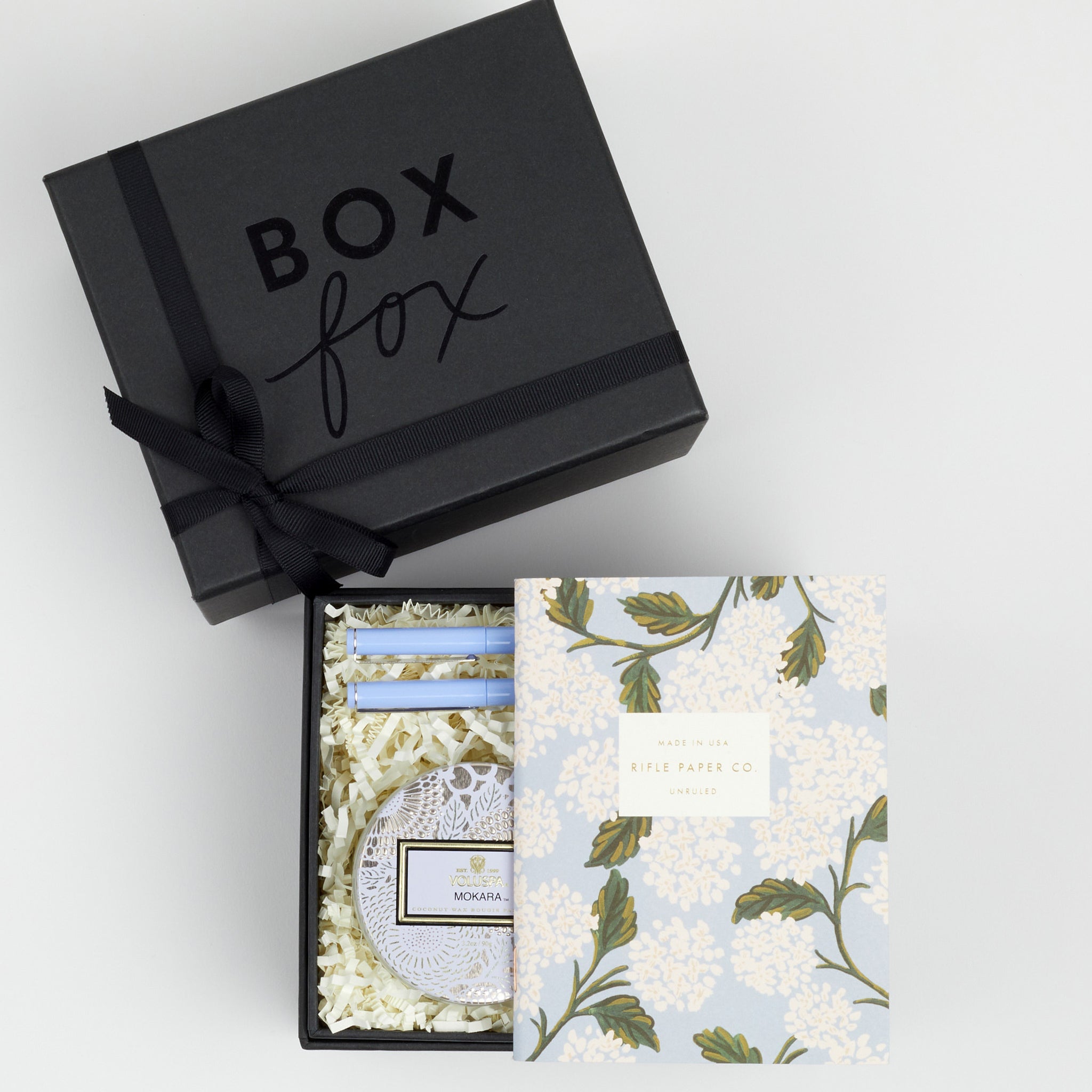 BOXFOX black Gift Box containing Rifle Paper Hydrangea Pocket Notebooks, 2 Periwinkle Le Pens, and Voluspa Mokara Petite Glass Jar Candle.
