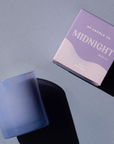 purple glass candle next to purple candle box behind light purple background