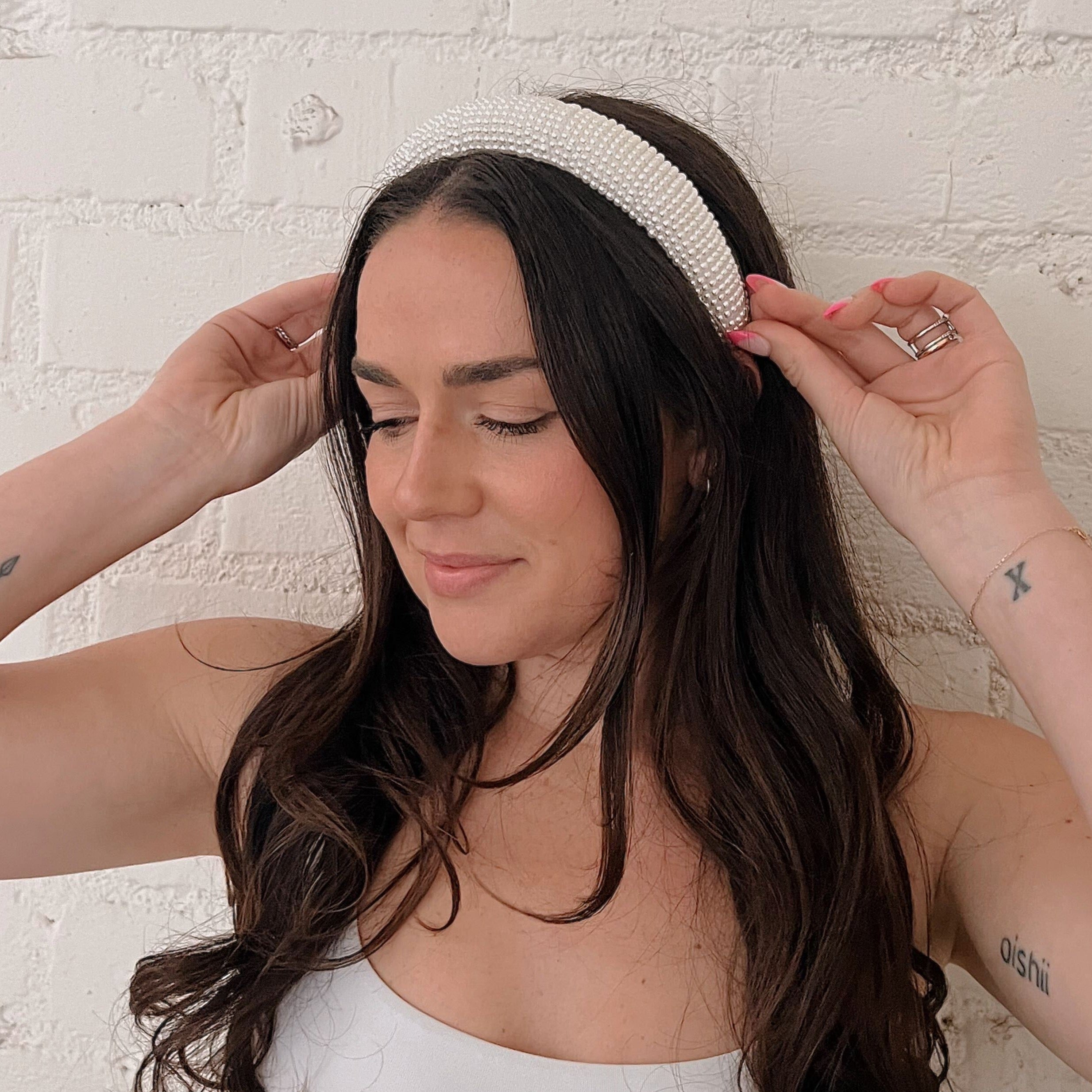 A woman with brown curly long hair wearing a white Diana pearl headband in her hair while she stands in front of a white brick background.