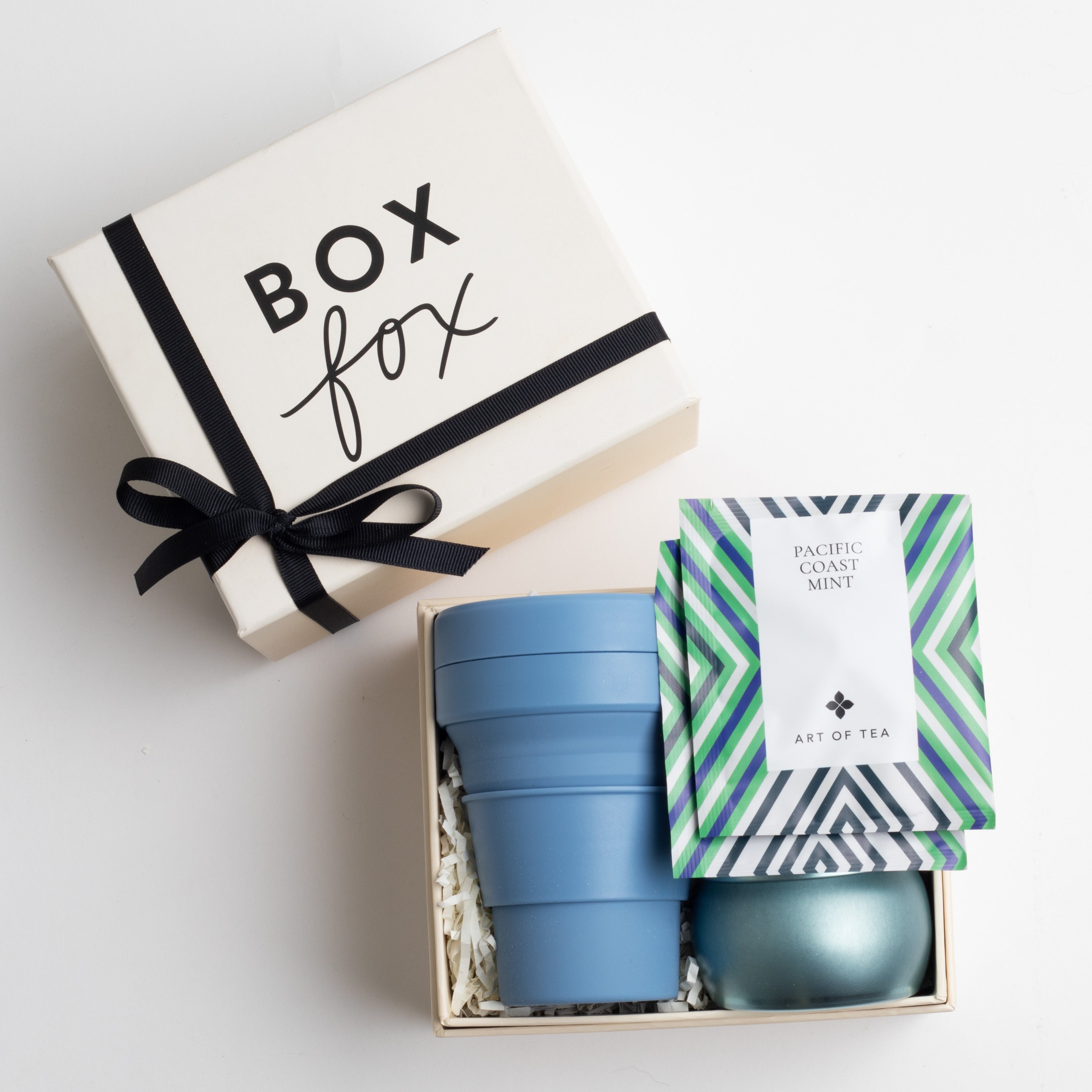 Creme BOXFOX gift box with a bow, next to an open gift box containing a blue collapsible cup, a blue tin candle and two art of tea sachets.